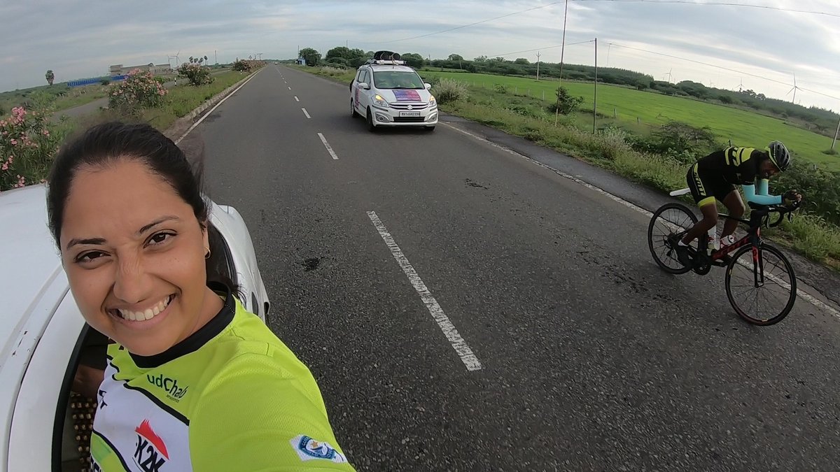 Jyoti Tripathi smiling from the crew vehicle window with Bharat Pannu riding behind her and the escort car ahead, on the Kashmir to Kanyakumari route